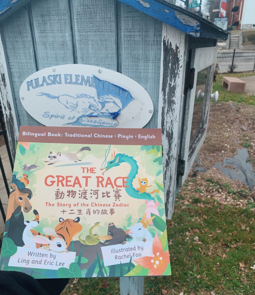 A person holds a book called The Great Race: The Story of the Chinese Zodiac in front of a little library at Pulaski Elementary.