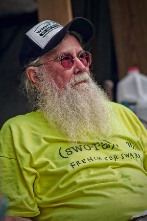 A photo of Jim webb wearing ayellow shirt. He is wearing a hat and glasses and has a long white beard.