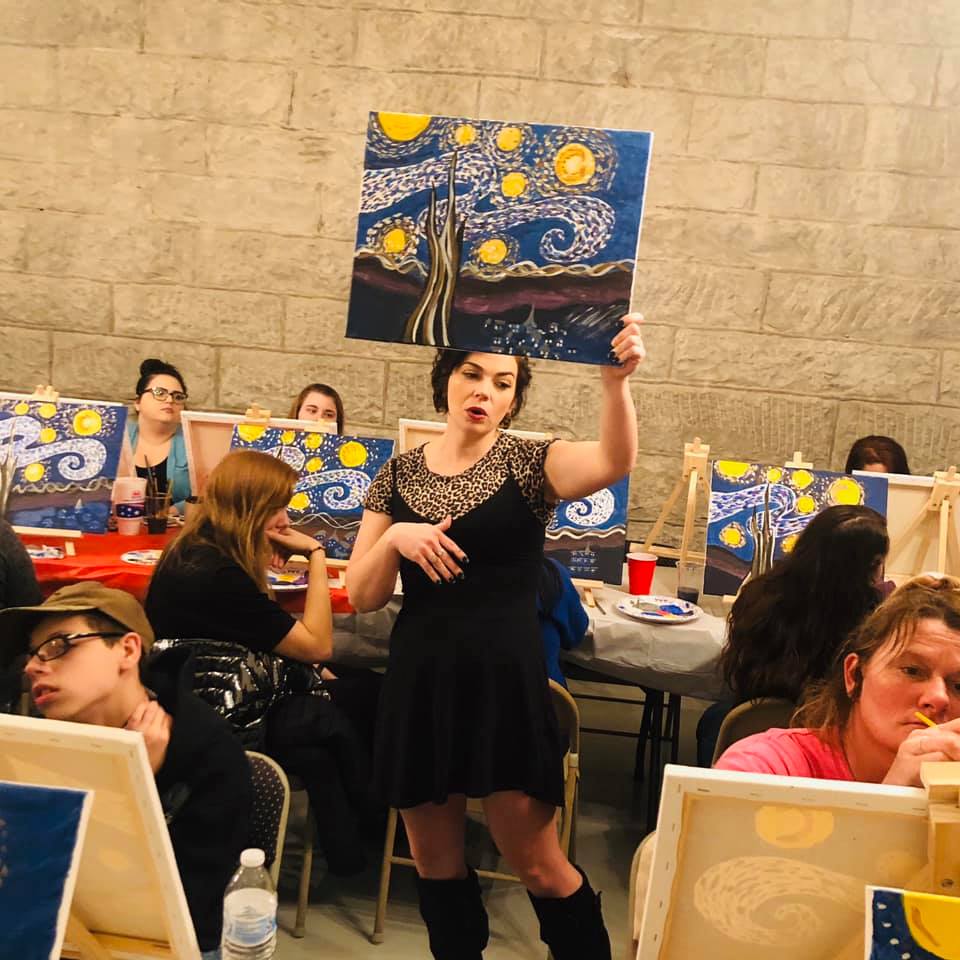 Art positive project director, jessica salyer, holds a painting above her head during a painting class. Multiple people are painting in the background.