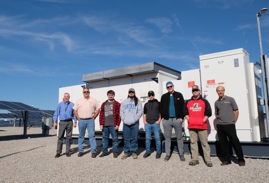 Several people stand in front of a big white inverter box and solar panels.