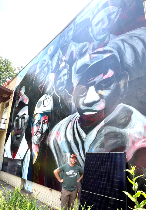 A mural of coal miners at Hemphill with Cameron standing in front of it next to a solar panel