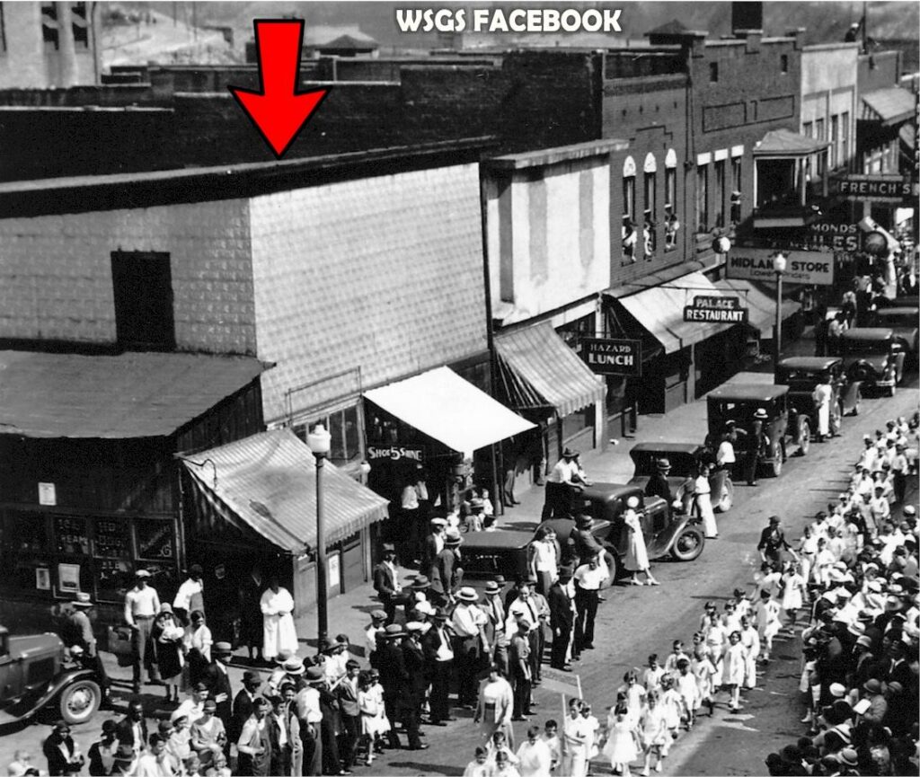 An old photo of main street with wooden buildings, old signs and awnings. The streets are crowded and their appears to be a parade.