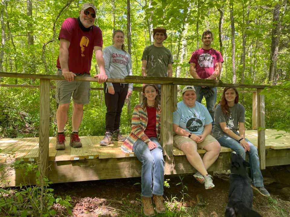 A group of university students sits on a foot bridge in the middle of a forest on berea college land