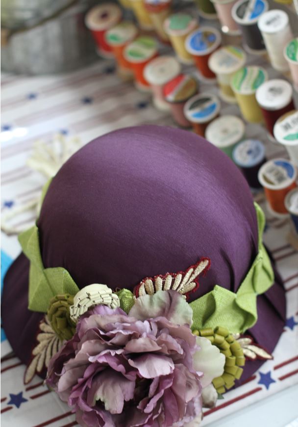 A purple hat sits on a table with spools of thread. The hat is made by glenna combs of sycamore hollow hats in manchester, kentucky
