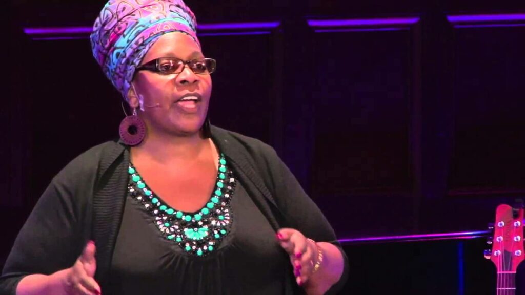 A Black person stands on stage delivering a TED talk