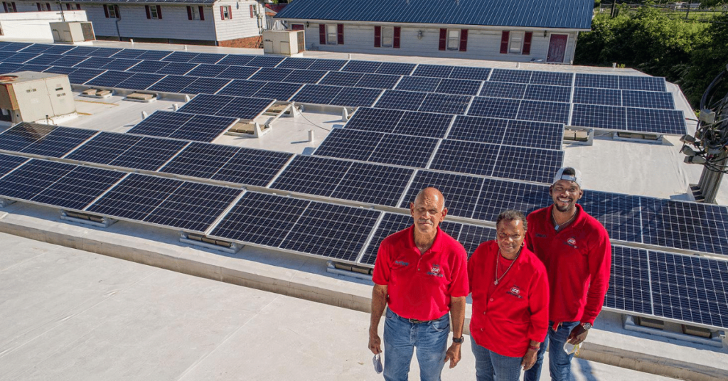 Three people in red shirts stand in front of solar panels on a roof in eastern kentucky.