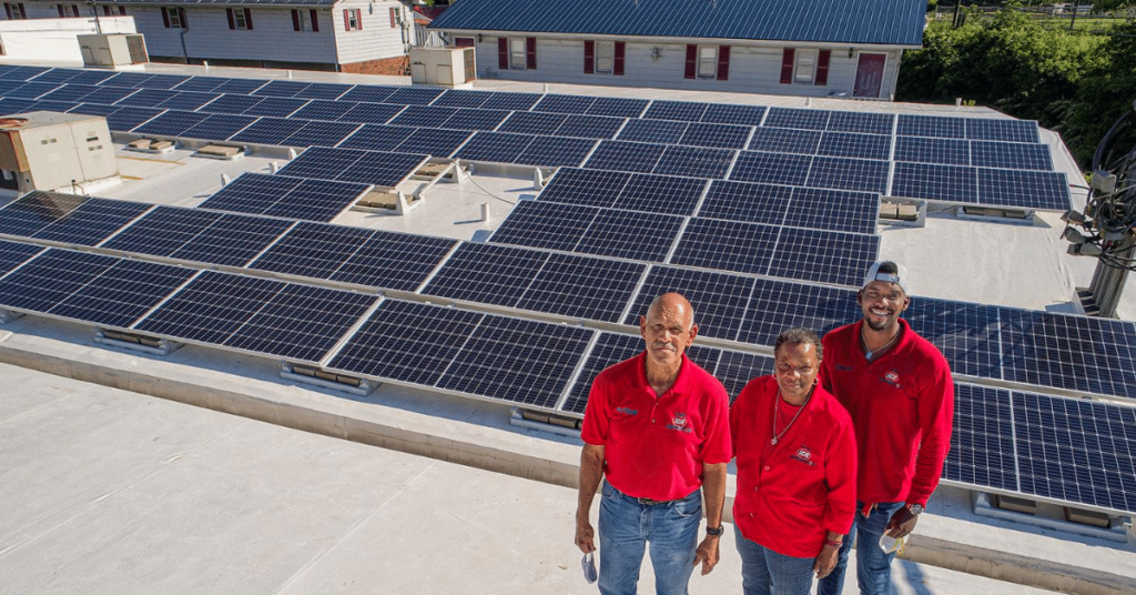 Three people stand on top of a roof next to several solar panels. The picture was taken at Isom IGA in letcher county.