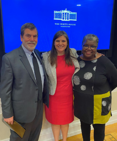 Three people stand in formal attire in front of a screen that says the white house