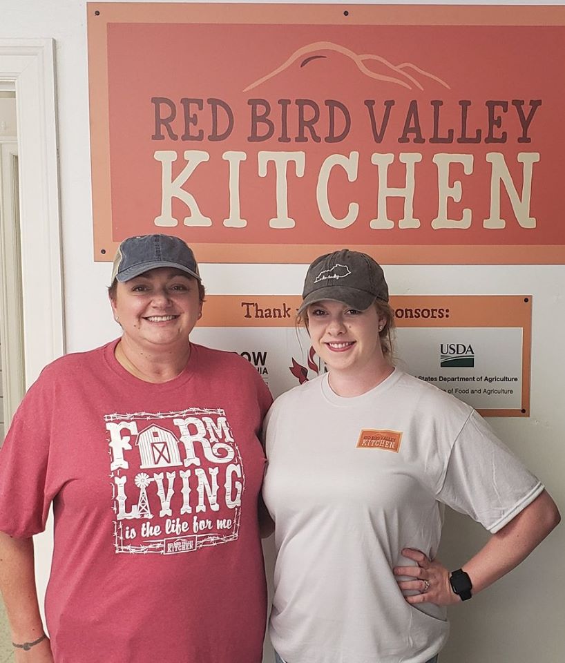 Two volunteers stand in front of a sign for Red Bird Mission's Red Bird Valley kitchen. Community members can use the kitchen to put up food.