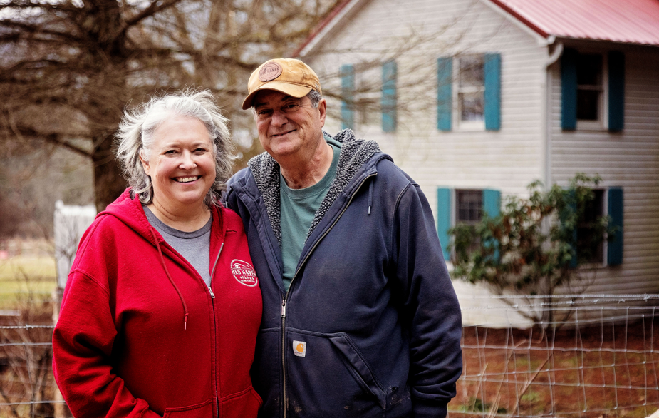 sandy and martin with red haven farm stand in front of the farmhouse that is available for rent and farm stays