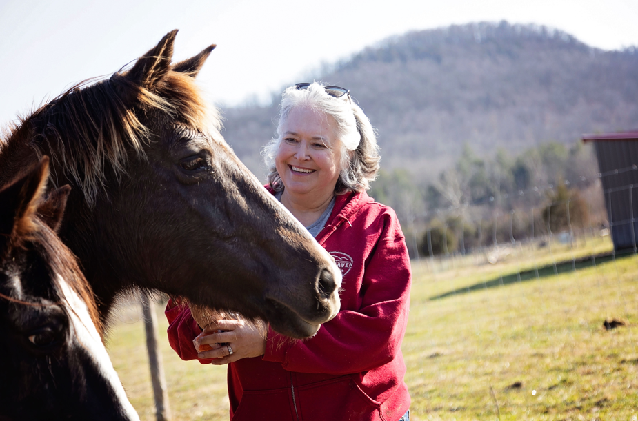 sandy with red haven farm stands with one of her horses with the hills of estill county in the background