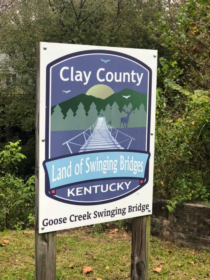 A sign that reads 'Clay County Land of Swinging Bridges Kentucky' stands in a field near a bridge. 