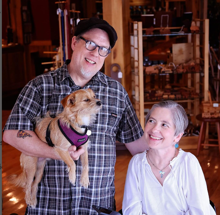 steve and heather johnson smiling at top drawer gallery. steve is holding stanley, a small brown dog