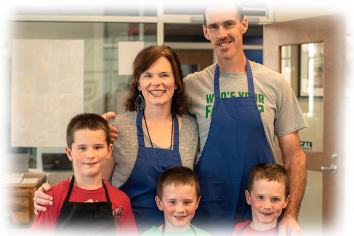 A family of five with two parents and three young kids stand looking at the camera. They are wearing aprons.