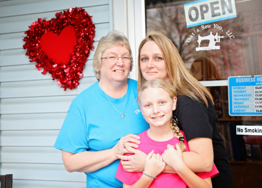 three people stand in front of their open sign hanging on the business door of thats sew you in stanton kentucky.