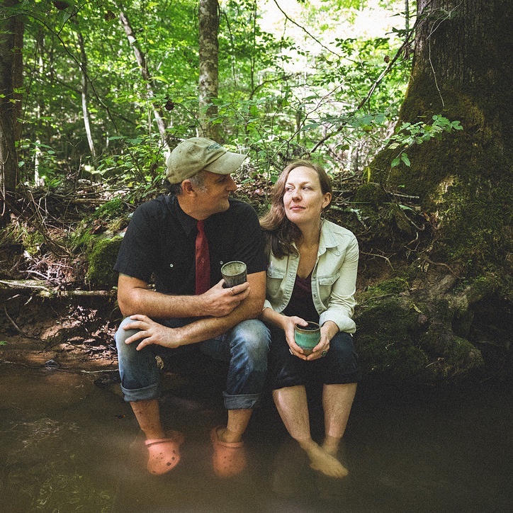 Casey and Laura sit with their feet in a creek drinking out of pottery
